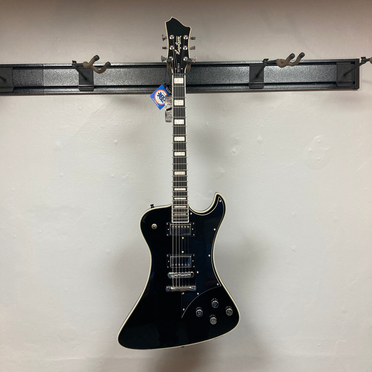 A Hagstrom Fantomen Custom Black w/case electric guitar, featuring a single cutaway, white binding, and dual Lundgren Design humbuckers, hangs on a wall-mounted guitar rack against a plain light-colored wall.