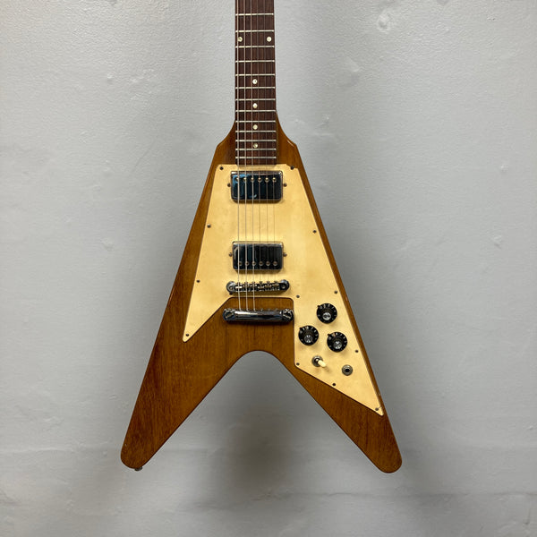 A brown 1975 Gibson Flying V electric guitar by Gibson, featuring a natural finish, distinctive V-shaped body, three control knobs, and two pickups, is displayed against a plain gray wall. Modeled after the classic vintage Flying V design, this instrument embodies the iconic style of its era and comes with a case.