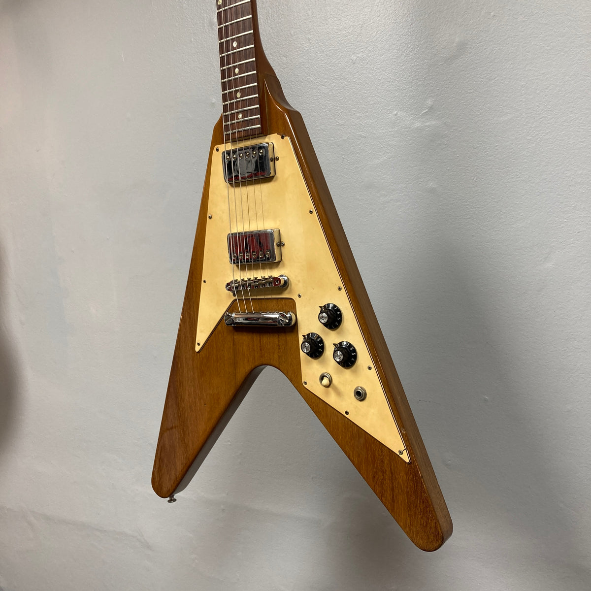 A 1975 Gibson Flying V electric guitar by Gibson, featuring a natural finish, V-shaped body, and cream pickguard, is mounted on a light gray wall.