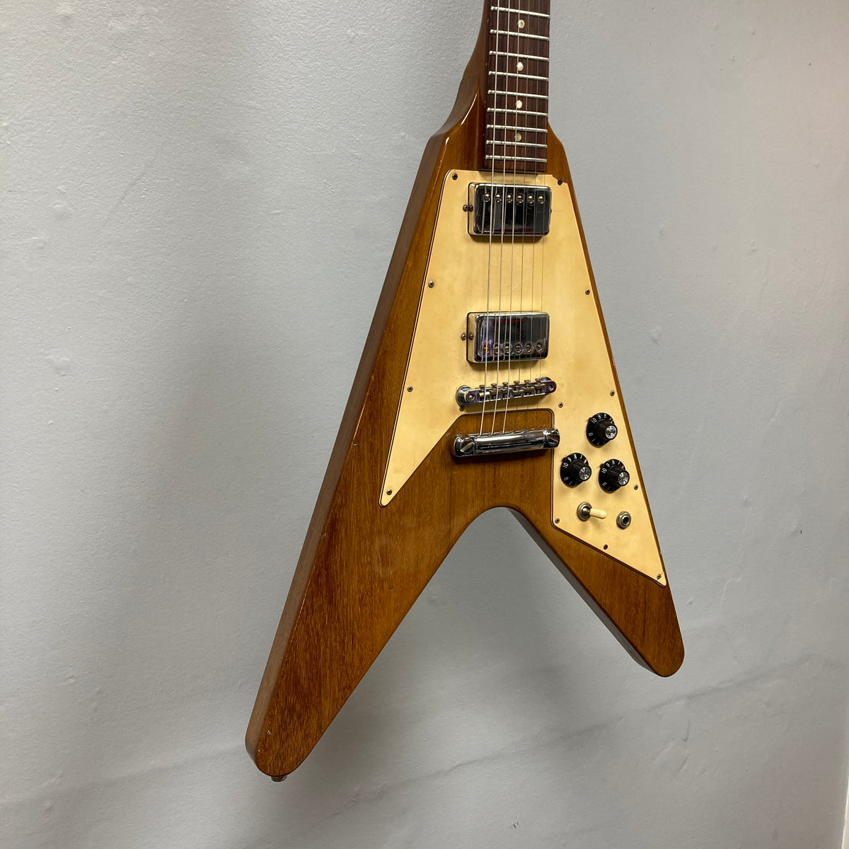 Close-up of a 1975 Gibson Flying V electric guitar with a natural finish, featuring a cream pickguard, two humbucker pickups, and control knobs, hanging against a plain light-colored wall. The iconic instrument comes with its original case.