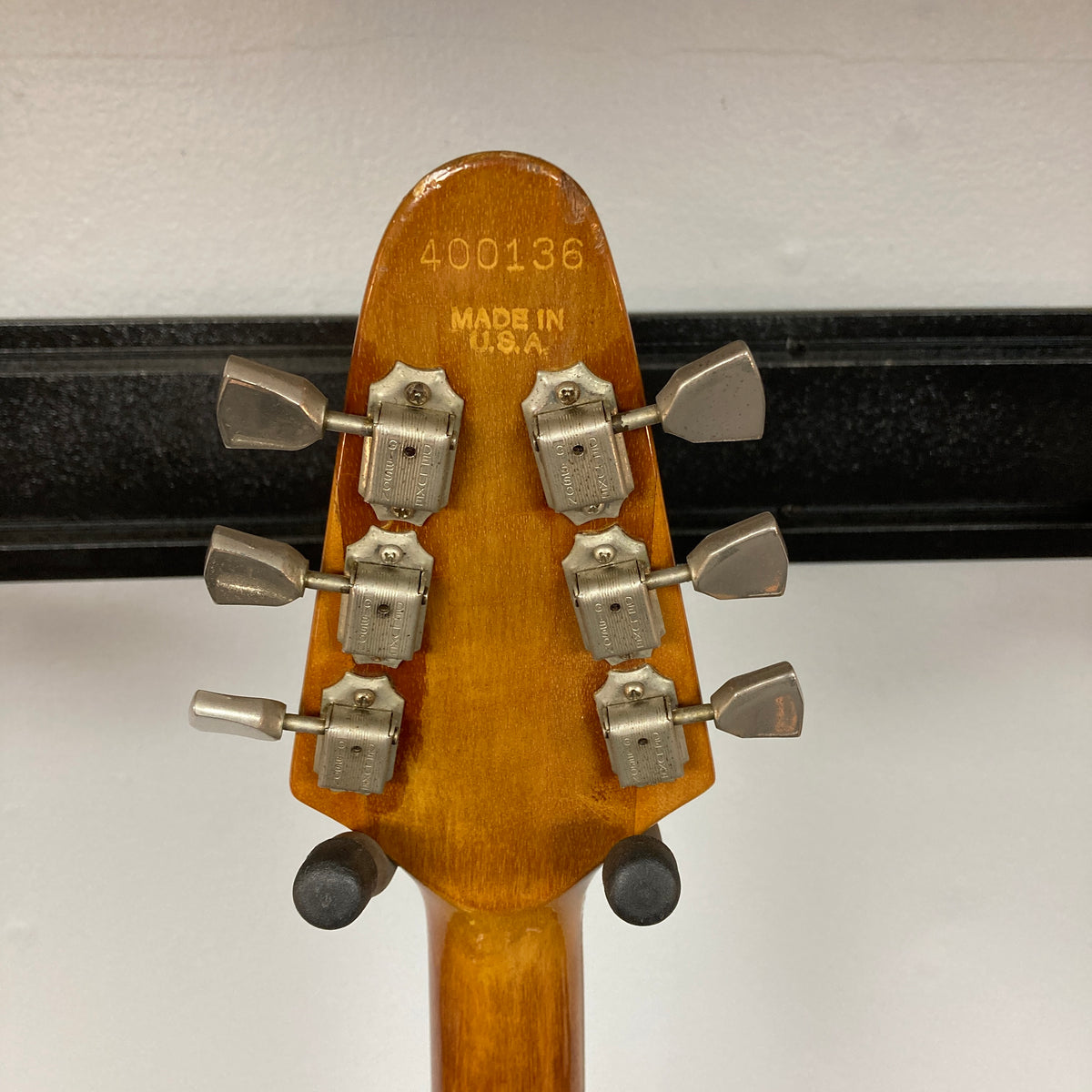 Close-up of the back of a Gibson 1975 Flying V w/Case headstock, displaying six tuning pegs, serial number 400136, and “Made in U.S.A.” engraved on the natural finish wood.