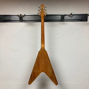 A 1975 Gibson Flying V w/Case by Gibson, featuring a natural finish, hangs by its headstock on a black wall-mounted rack, displayed from the back against a white wall.