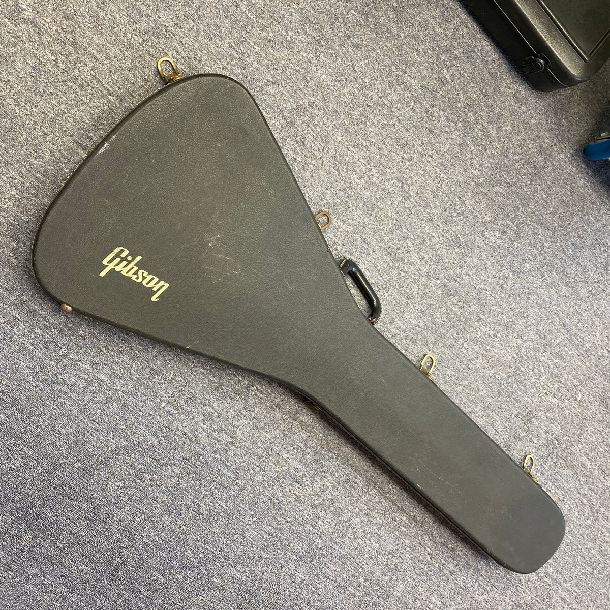 A black, hard-shell Gibson case featuring a gold Gibson logo—designed to fit the 1975 Gibson Flying V w/Case or other vintage Flying V models—is lying closed on a gray carpeted floor.