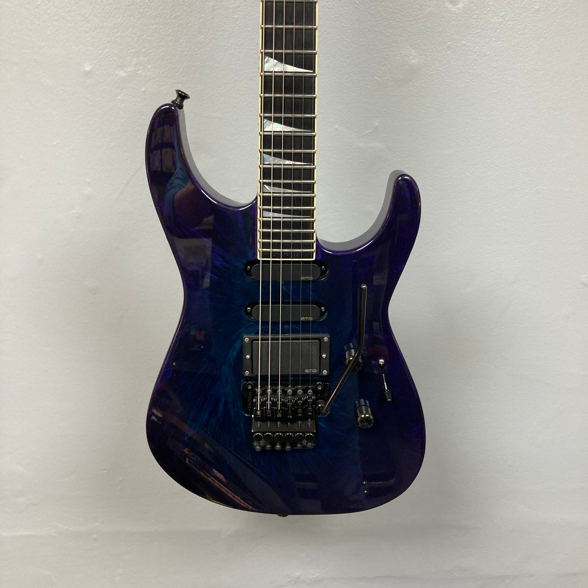A 1995 Jackson SL1 Soloist Custom Shop Eerie Dess Swirl electric guitar by Jackson, featuring a striking blue double-cutaway body, three pickups, a Floyd Rose bridge, and sleek black hardware, hangs against a plain white wall.