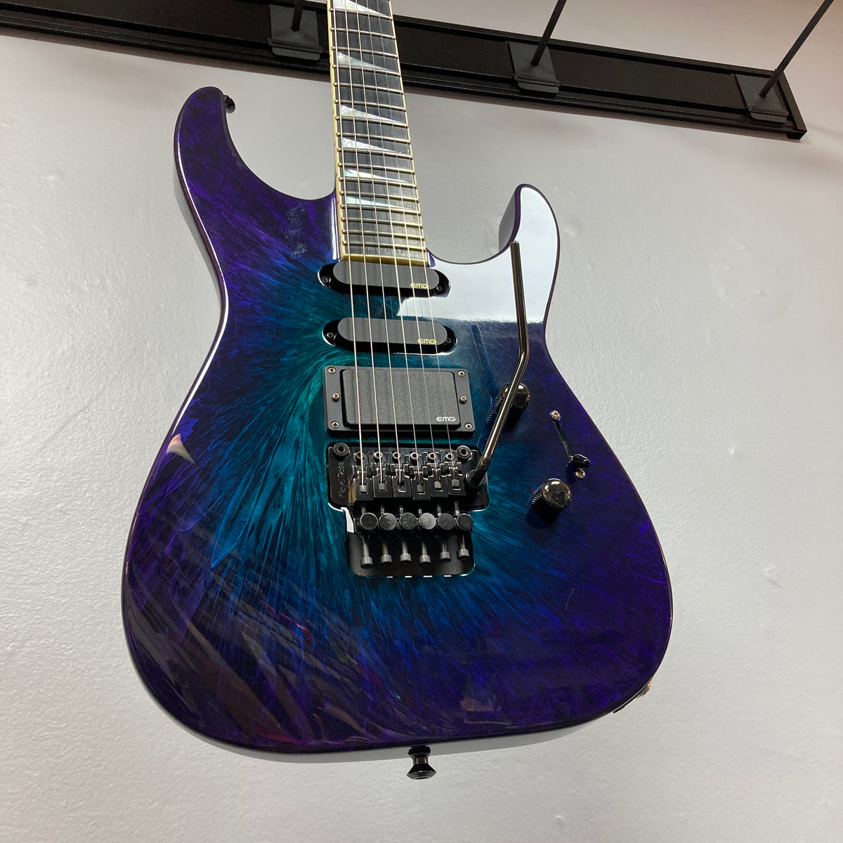 Hanging on the wall is a 1995 Jackson SL1 Soloist Custom Shop Eerie Dess Swirl electric guitar, featuring a striking purple and blue burst finish, black pickups, a Floyd Rose tremolo bridge, and a sleek maple neck—an iconic model from Jackson’s Custom Shop collection.