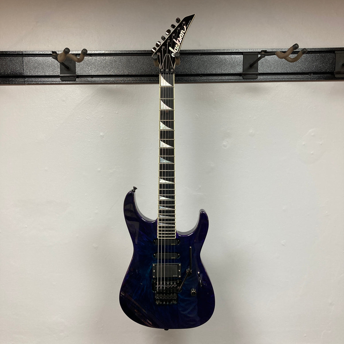 A 1995 Jackson SL1 Soloist Custom Shop Eerie Dess Swirl electric guitar by Jackson, featuring a pointed headstock and triangular inlays, is displayed on a wall-mounted guitar rack against a plain white wall.
