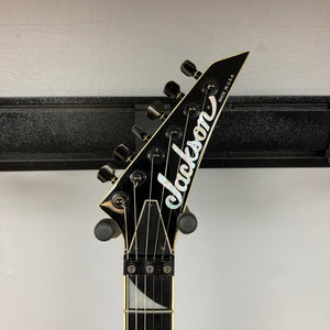Close-up of the headstock of a black 1995 Jackson SL1 Soloist Custom Shop Eerie Dess Swirl electric guitar, featuring six tuning pegs, the Jackson logo, and “Made in U.S.A.” text, mounted on a wall hanger against a white background.