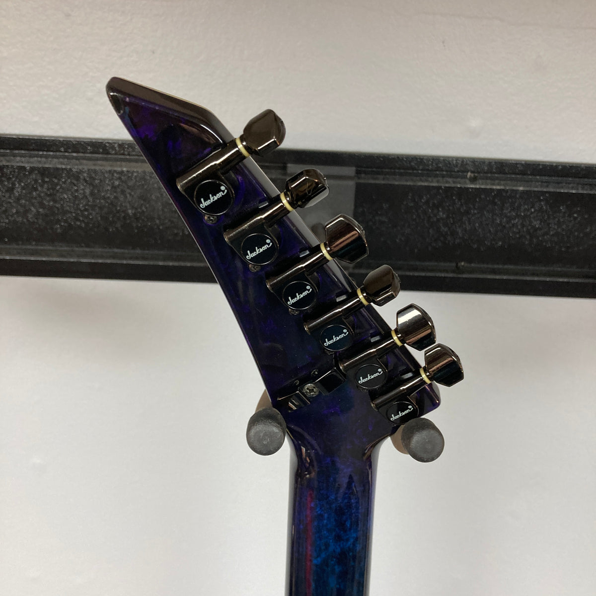 Close-up of the back of a dark electric guitar’s headstock with six Jackson-branded tuning pegs, hanging on a wall-mounted holder—this striking piece is a 1995 Jackson SL1 Soloist Custom Shop Eerie Dess Swirl w/Case.