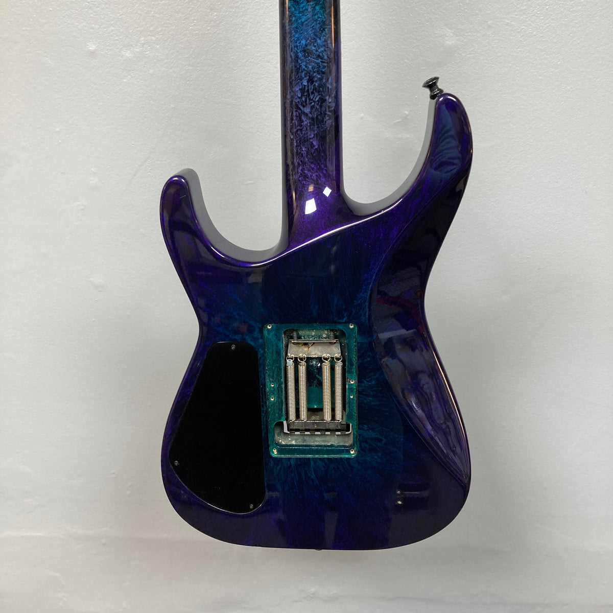 Rear view of a 1995 Jackson SL1 Soloist Custom Shop Eerie Dess Swirl electric guitar, featuring a glossy blue and purple marbled finish, exposed electronics cavity with no back cover plate, made by Jackson’s USA Custom Shop, and positioned against a plain white wall.