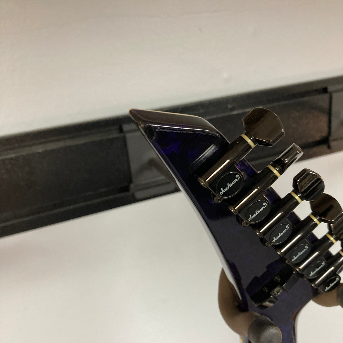 Close-up of the headstock of a Jackson 1995 SL1 Soloist Custom Shop Eerie Dess Swirl electric guitar, featuring black tuning pegs, mounted on a wall hanger against a white wall.