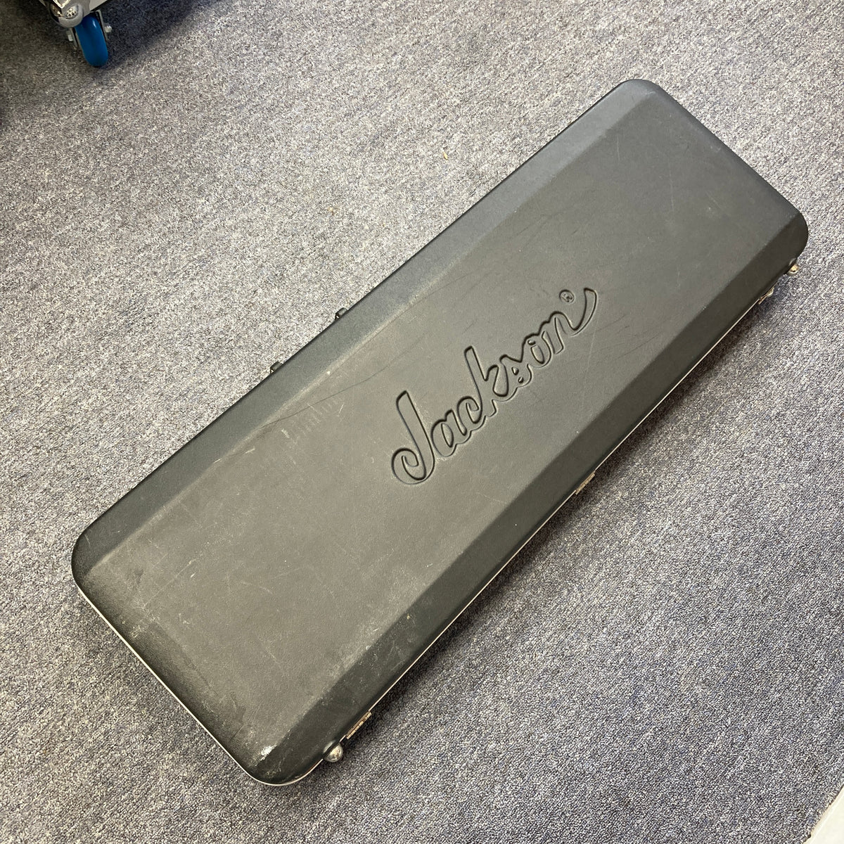 A black Jackson hard guitar case, featuring the “Jackson” logo and made for the 1995 Jackson SL1 Soloist Custom Shop Eerie Dess Swirl w/Case, is lying closed on a gray carpeted floor.