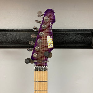 A close-up of the headstock of a 2019 ESP USA M-II Purple Haze Burst electric guitar reveals six tuning pegs, string guides, and part of the wooden fretboard, all showcased in a striking Purple Haze Burst finish against a white wall. This American-made ESP guitar exemplifies exceptional craftsmanship and an eye-catching design, and comes complete with its original case.