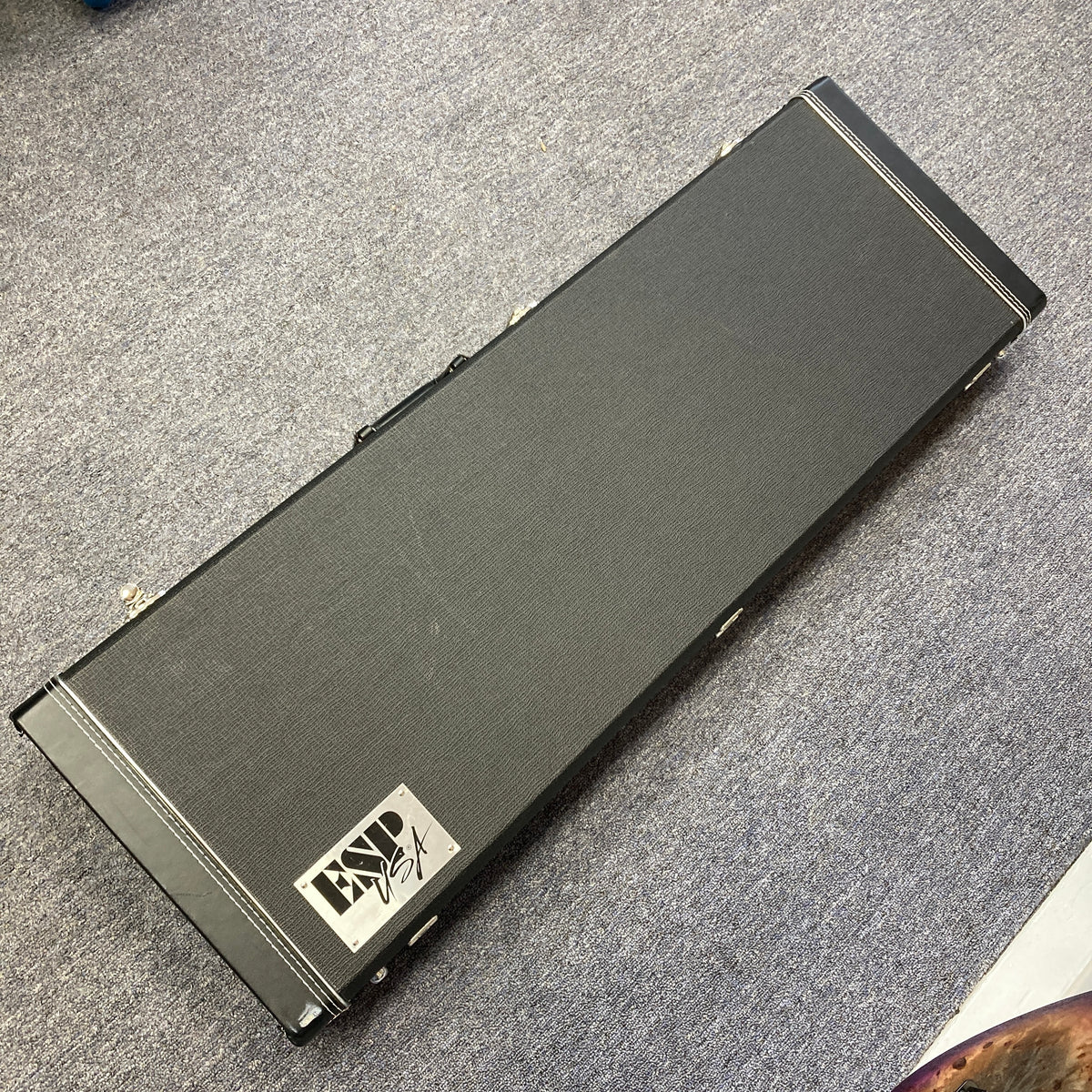 A black rectangular hard case with a handle, featuring a “Lynx USA” label, rests on a gray carpeted floor—perfect for safely storing your ESP 2019 USA M-II Purple Haze Burst electric guitar or proudly presenting this exceptional American-made ESP model.
