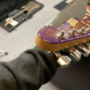 Close-up of the headstock and tuning pegs of a 2019 ESP USA M-II in striking Purple Haze Burst finish, highlighting one of ESP’s premier American-made guitars, with part of a person’s arm and the included case visible in the background.