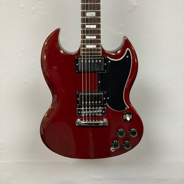A 1977 Gibson SG Standard Cherry electric guitar by Gibson, featuring a classic double-cutaway mahogany body, two humbucker pickups, four control knobs, and a black pickguard evocative of the iconic Kalamazoo era, hangs elegantly against a plain white wall.