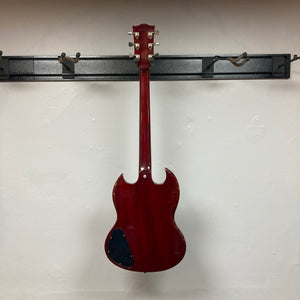 A 1977 Gibson SG Standard Cherry electric guitar by Gibson hangs on a wall-mounted rack, displayed from the back to reveal its visible tuning pegs and a black patch on the lower mahogany body.