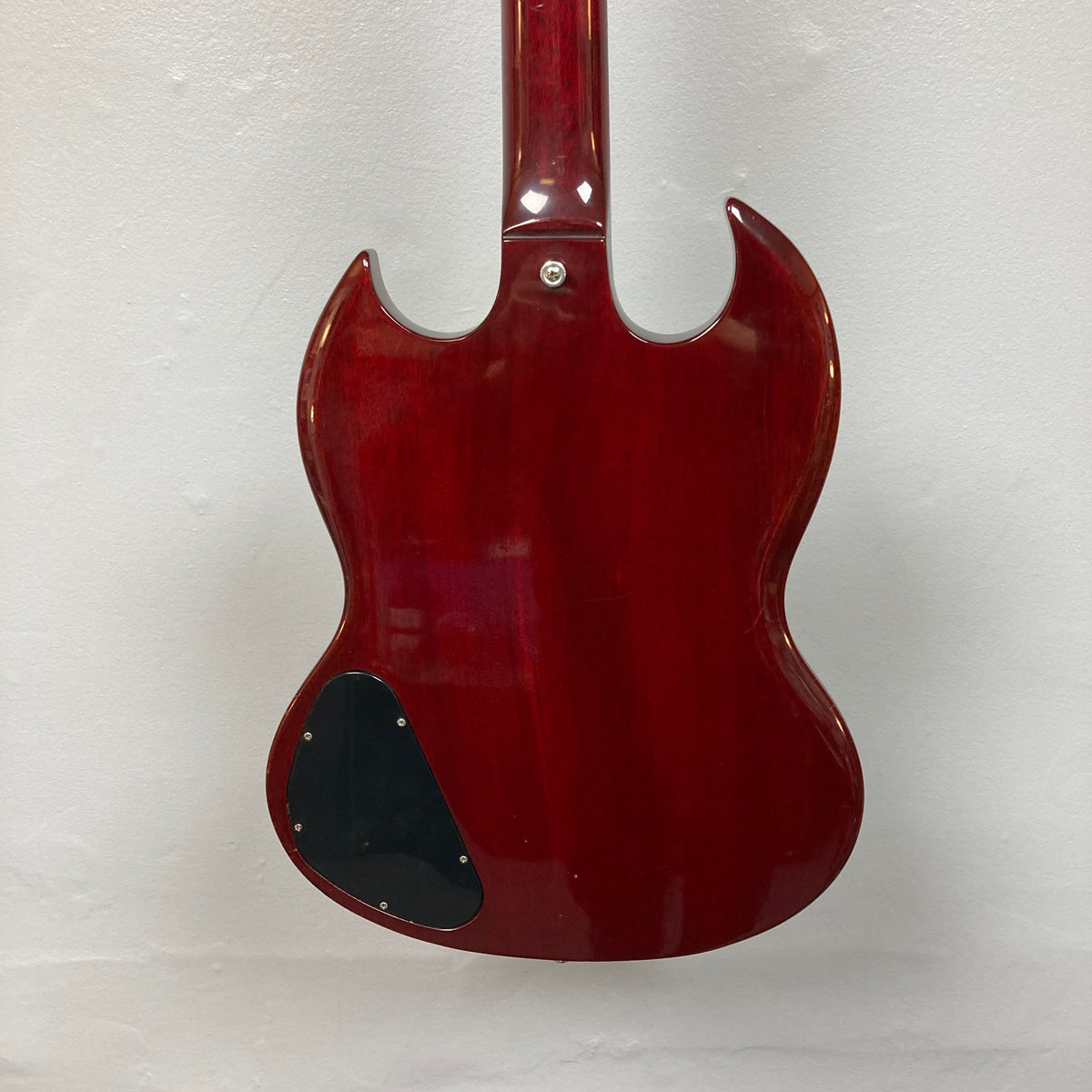 The back of a 1977 Gibson SG Standard Cherry electric guitar by Gibson is shown against a white wall, highlighting its double cutaway mahogany body. A black plastic control cover appears on the lower left side, echoing the classic style of the Gibson Kalamazoo era.