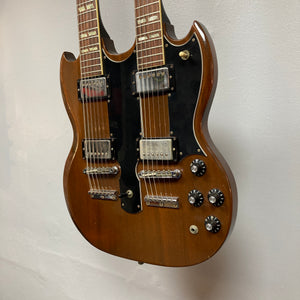 A 1978 Gibson EDS-1275 Double Neck Walnut w/Case by Gibson—featuring a rich brown walnut finish, black pickguard, four pickups, four control knobs, and metallic hardware—hangs against a plain wall, exemplifying style and versatility.
