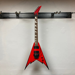A Jackson Phil Demmel Demmelition King V electric guitar, featuring a striking red jagged V-shaped body and black hardware, hangs on a wall-mounted rack against a white background, showcasing the aggressive styling of this Jackson signature model.
