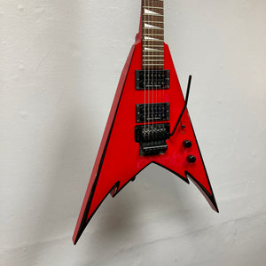A Jackson Phil Demmel Demmelition King V electric guitar by Jackson, featuring a red pointed V-shaped body with black trim, dual humbucker pickups, and a Floyd Rose tremolo whammy bar, photographed against a plain white wall.