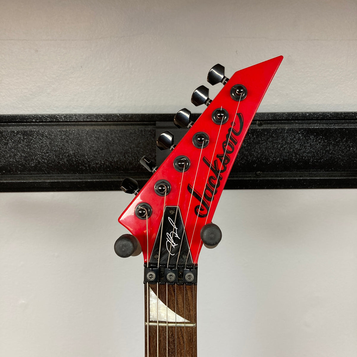 Close-up of the headstock of a red Jackson Phil Demmel Demmelition King V w/Case guitar by Jackson, featuring black tuning pegs, a signature, Floyd Rose tremolo, and the “Jackson” logo against a white wall background.