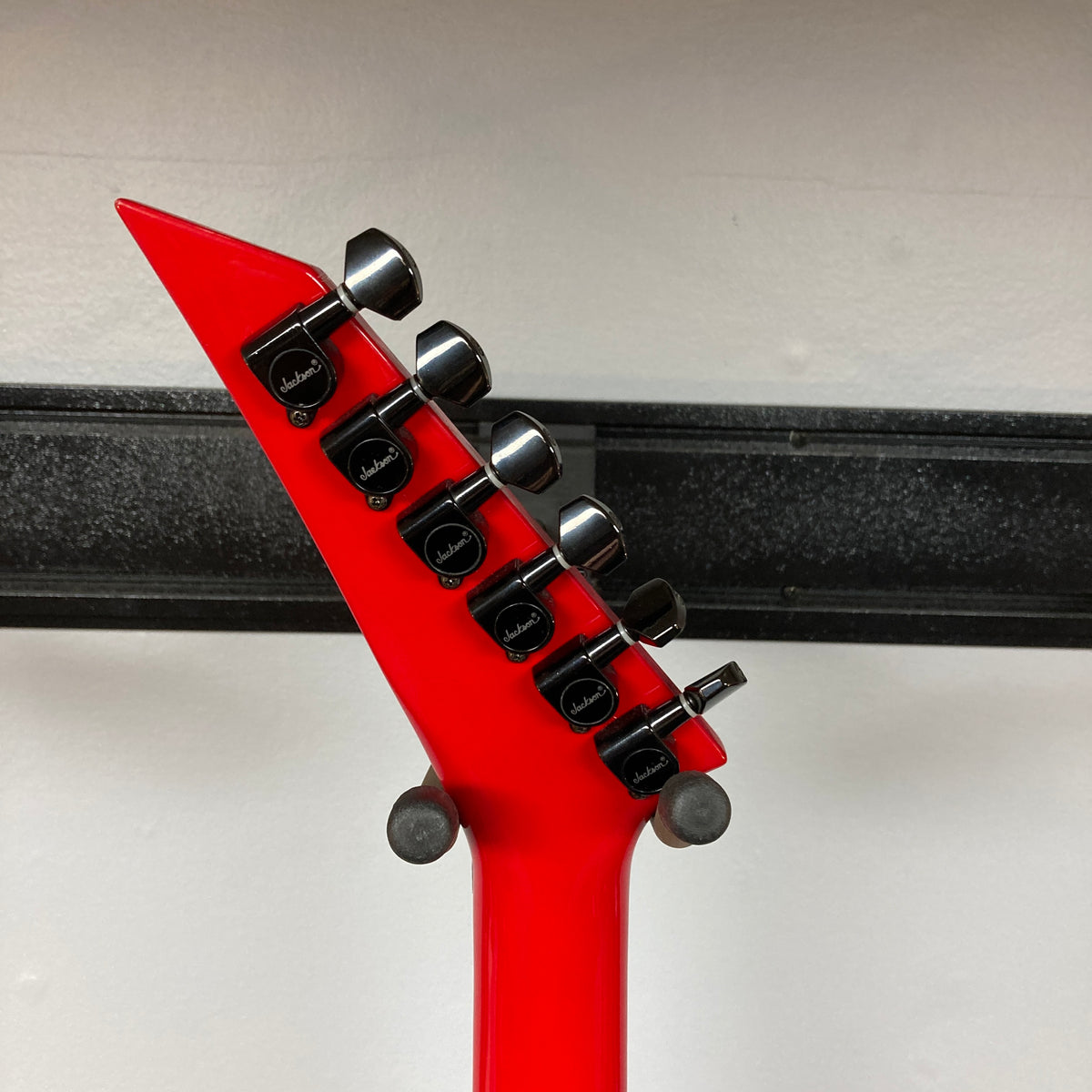 Close-up of the headstock and tuning pegs on a red Jackson Phil Demmel Demmelition King V w/Case electric guitar by Jackson, photographed against a white wall with a black shelf in the background.
