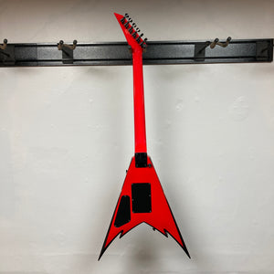 A bold Jackson Phil Demmel Demmelition King V w/Case by Jackson, featuring a red pointed V-shaped body with black accents and a sleek Floyd Rose tremolo system, hangs upright on a wall rack—its distinctive signature design making it instantly eye-catching.