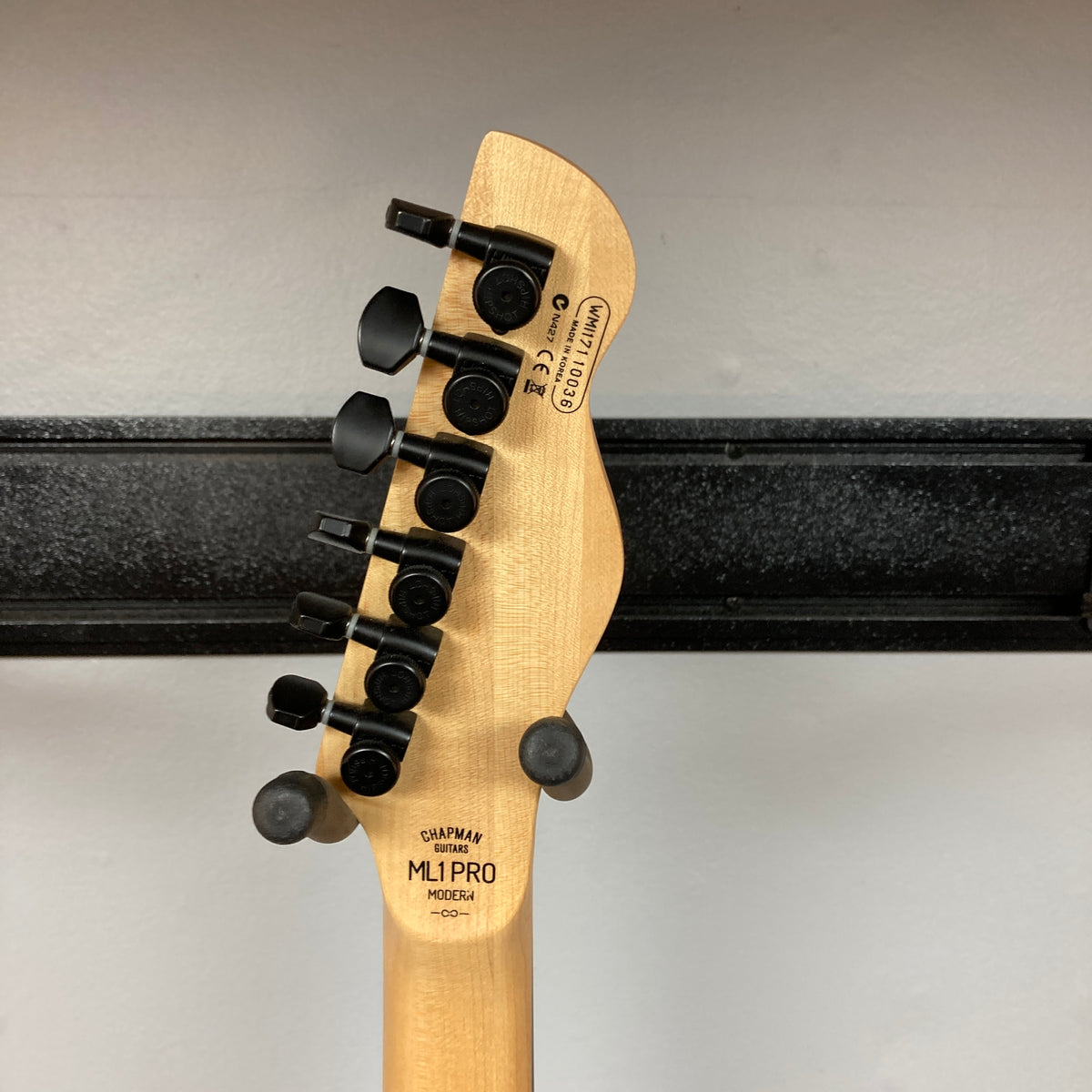The back of a Chapman ML1 Pro Modern Lunar electric guitar headstock with six black tuning pegs, displaying model and serial number details, photographed against a white wall and black horizontal rack.