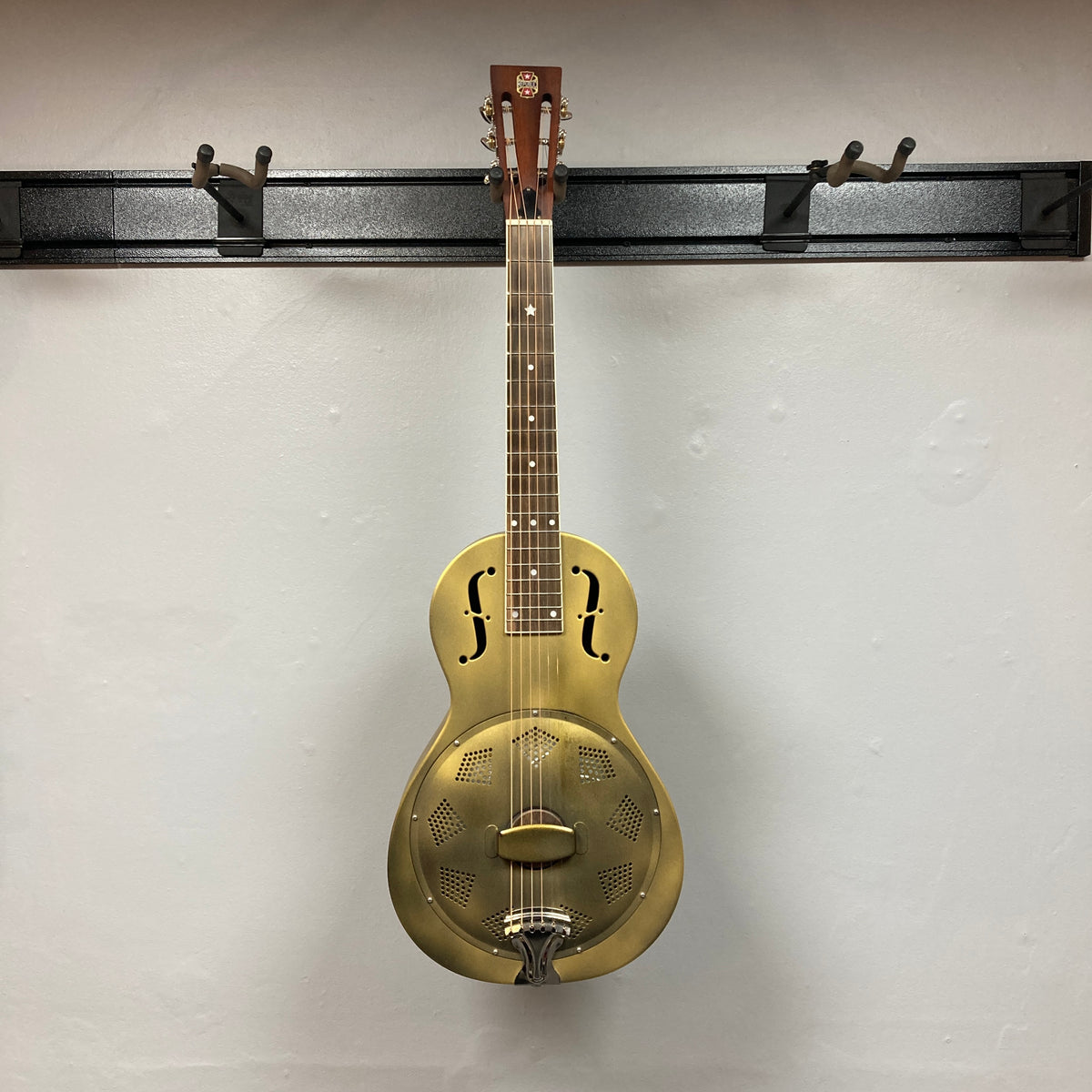 The Republic Parlor Resolian Antique Bronze Resonator w/Case by Republic, featuring a metal body with f-holes and a single cone, hangs on a wall-mounted rack against a white background, showcasing its subtle antique bronze finish.