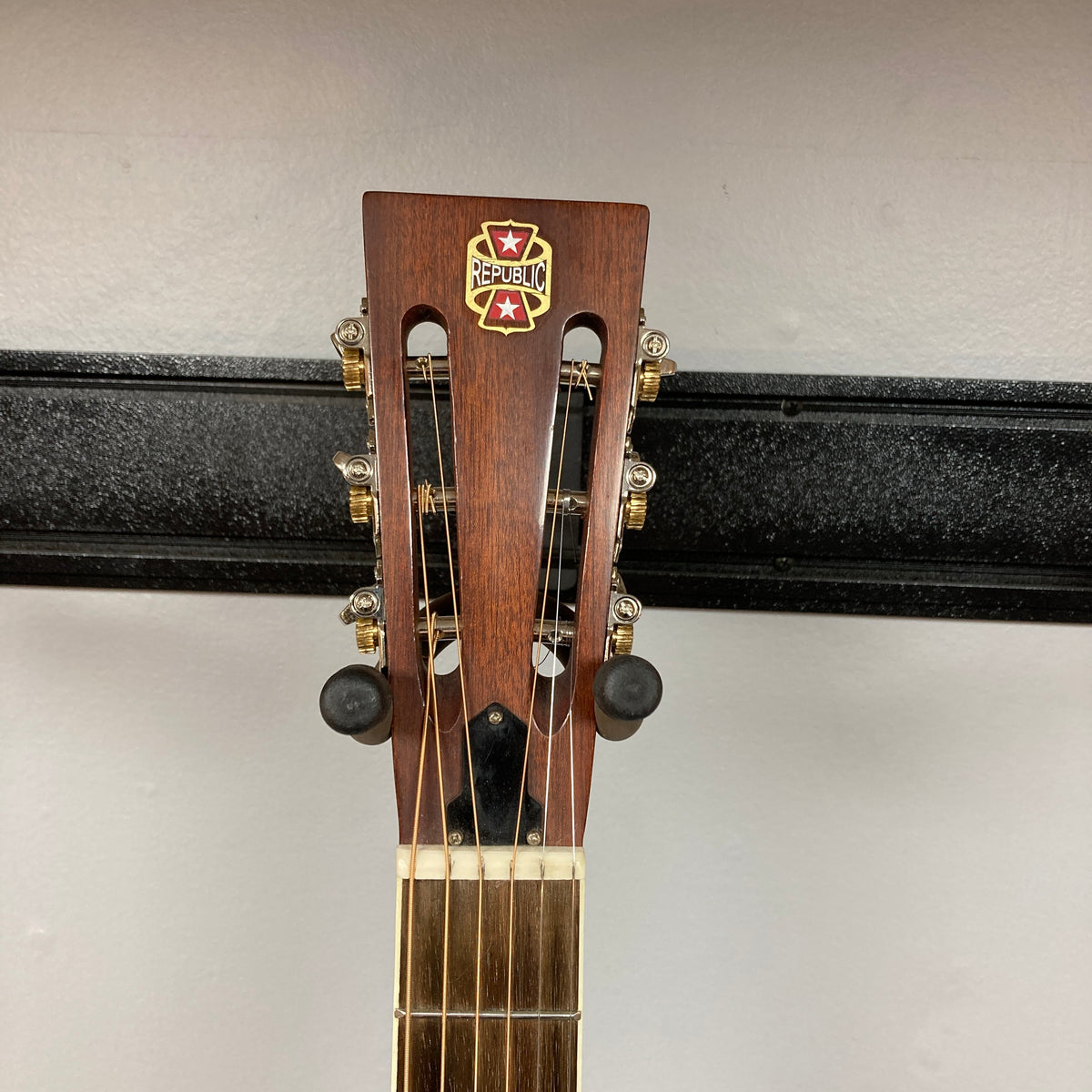 Close-up of the Republic Parlor Resolian Antique Bronze Resonator headstock, featuring the Republic logo, six tuning pegs, and a light-colored fretboard. Its single cone design gleams through the antique bronze finish, set against a plain wall and black rack.