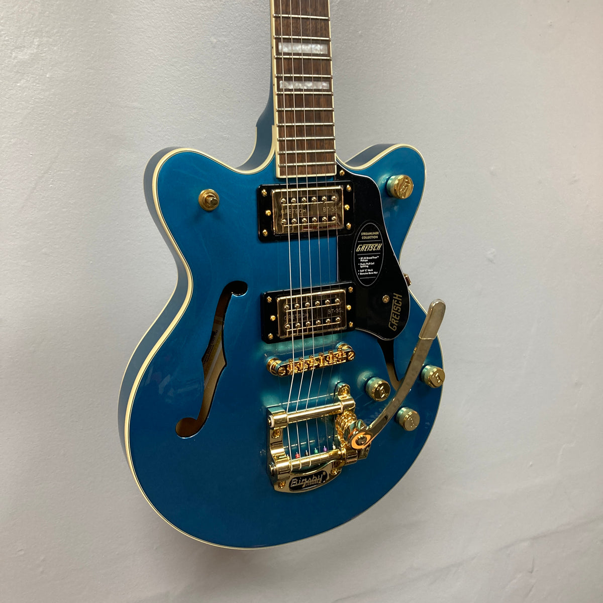 A Gretsch G2655TG Streamliner Riviera Blue Refurb semi-hollow electric guitar with gold hardware, Broad’Tron BT 3S pickups, a Bigsby tremolo, and an f-hole is hanging on a white wall.