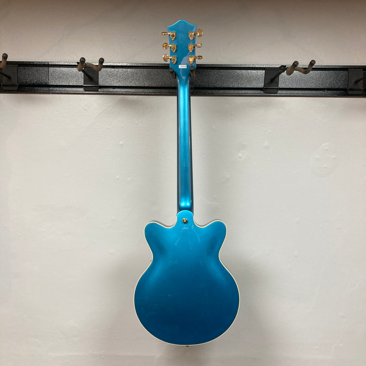 A Gretsch G2655TG Streamliner Riviera Blue Refurb semi-hollow electric guitar is seen from the back as it hangs on a wall-mounted rack with empty hooks against a plain white wall.