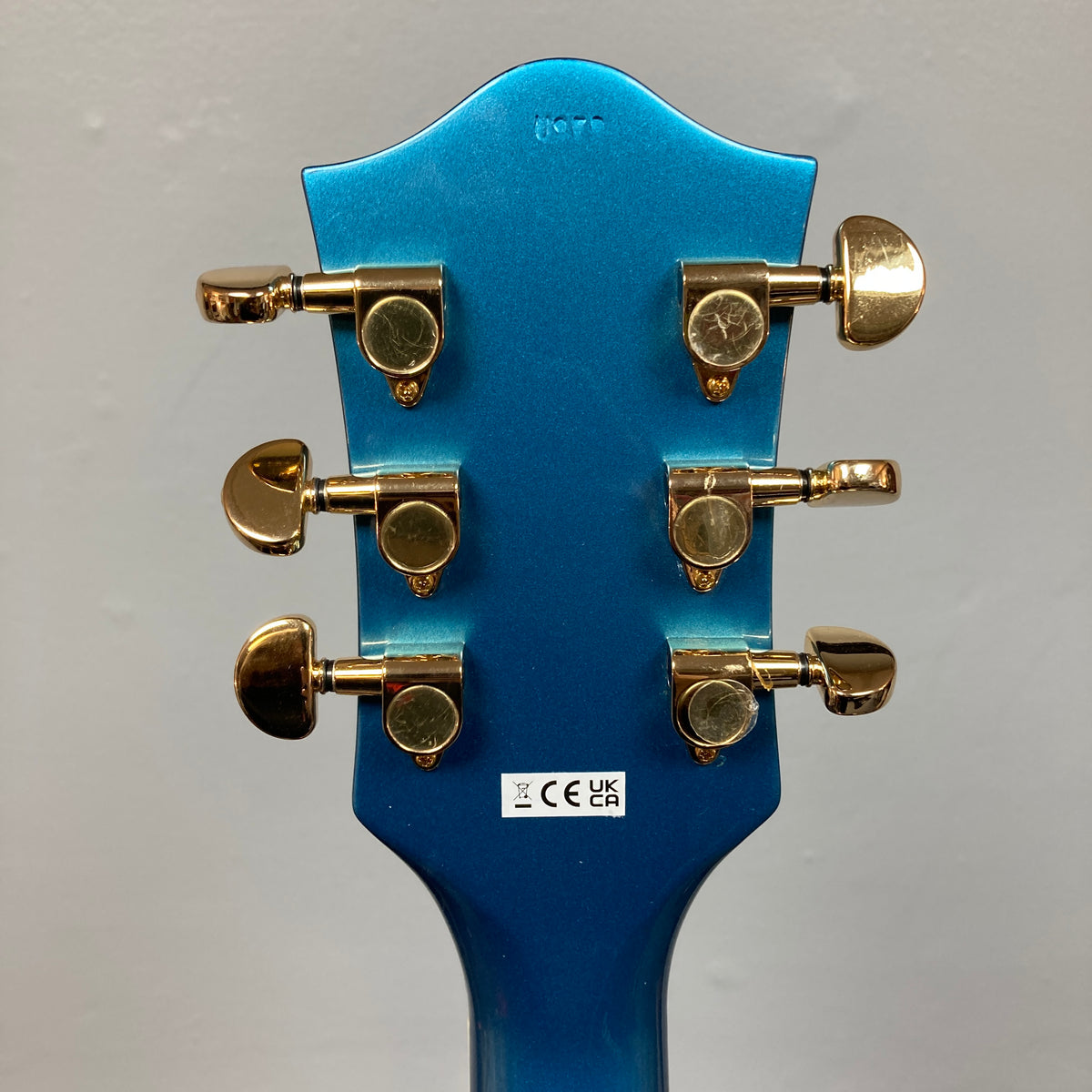 Back of a Riviera Blue Gretsch G2655TG Streamliner Refurb semi-hollow electric guitar headstock featuring six gold tuning pegs and a white sticker with certification symbols.