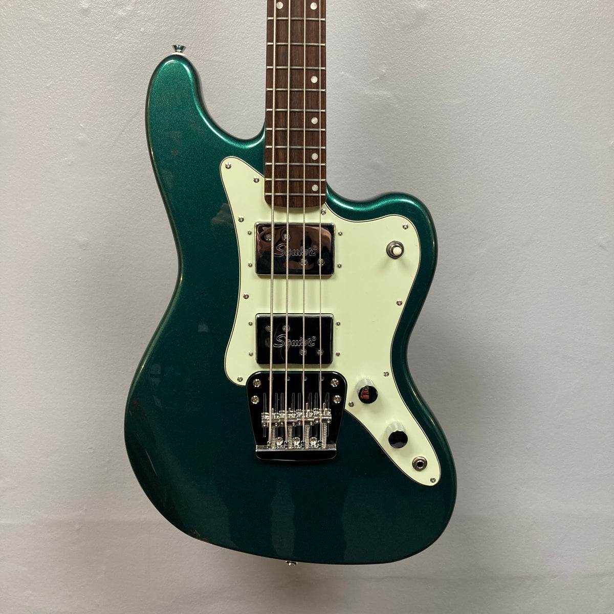 A Squier Paranormal Rascal Bass HH Sherwood Green Refurb, featuring a white pickguard, Fender Designed Wide Range humbuckers, four control knobs, and a chrome bridge, stands against a plain light gray wall.