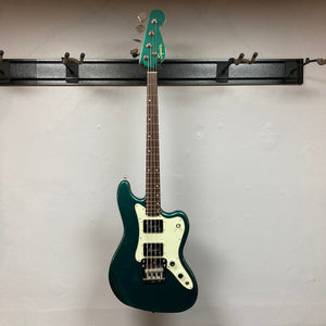 A Sherwood Green Squier Paranormal Rascal Bass HH is hanging on a black wall-mounted rack against a plain light-colored wall, its eye-catching finish and Fender-designed Wide Range humbuckers capturing the vibrant style for which Squier is known.