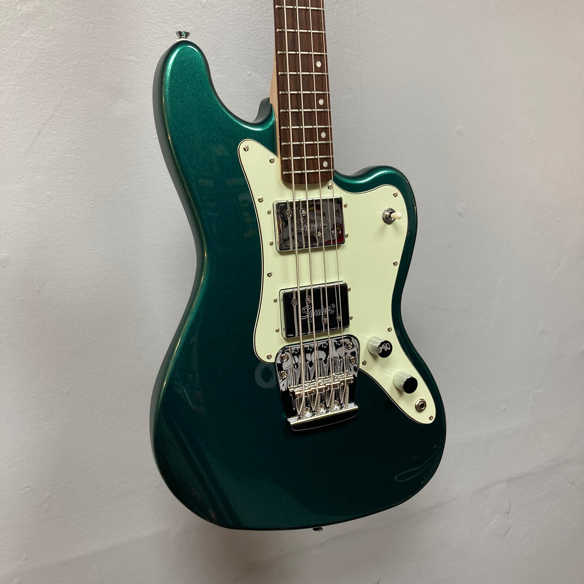 A Squier Paranormal Rascal Bass HH in Sherwood Green with a white pickguard, two pickups, and a chrome bridge hangs on a plain light-colored wall.