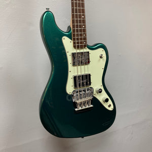 A Squier Paranormal Rascal Bass HH in Sherwood Green with a white pickguard, two pickups, and a chrome bridge hangs on a plain light-colored wall.