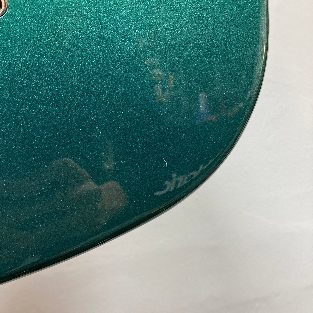 Close-up of the green metallic surface of a Squier Paranormal Rascal Bass HH Sherwood Green Refurb by Squier, highlighting a small white scratch near the edge and faint reflections.