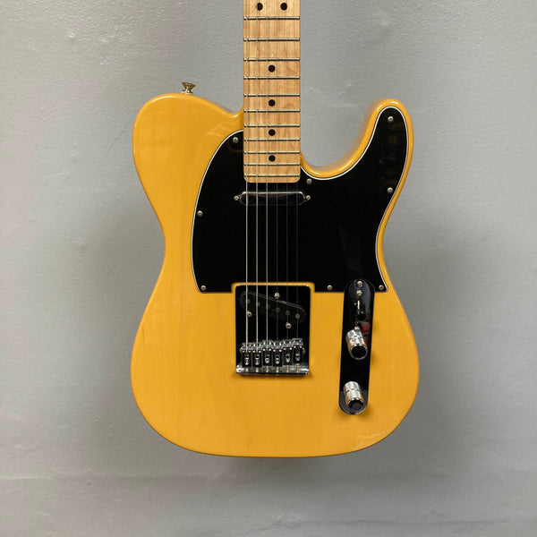 A Fender Player Telecaster 75th Anniversary Butterscotch Used electric guitar by Fender, featuring a black pickguard and maple fingerboard, is hanging against a light gray wall.