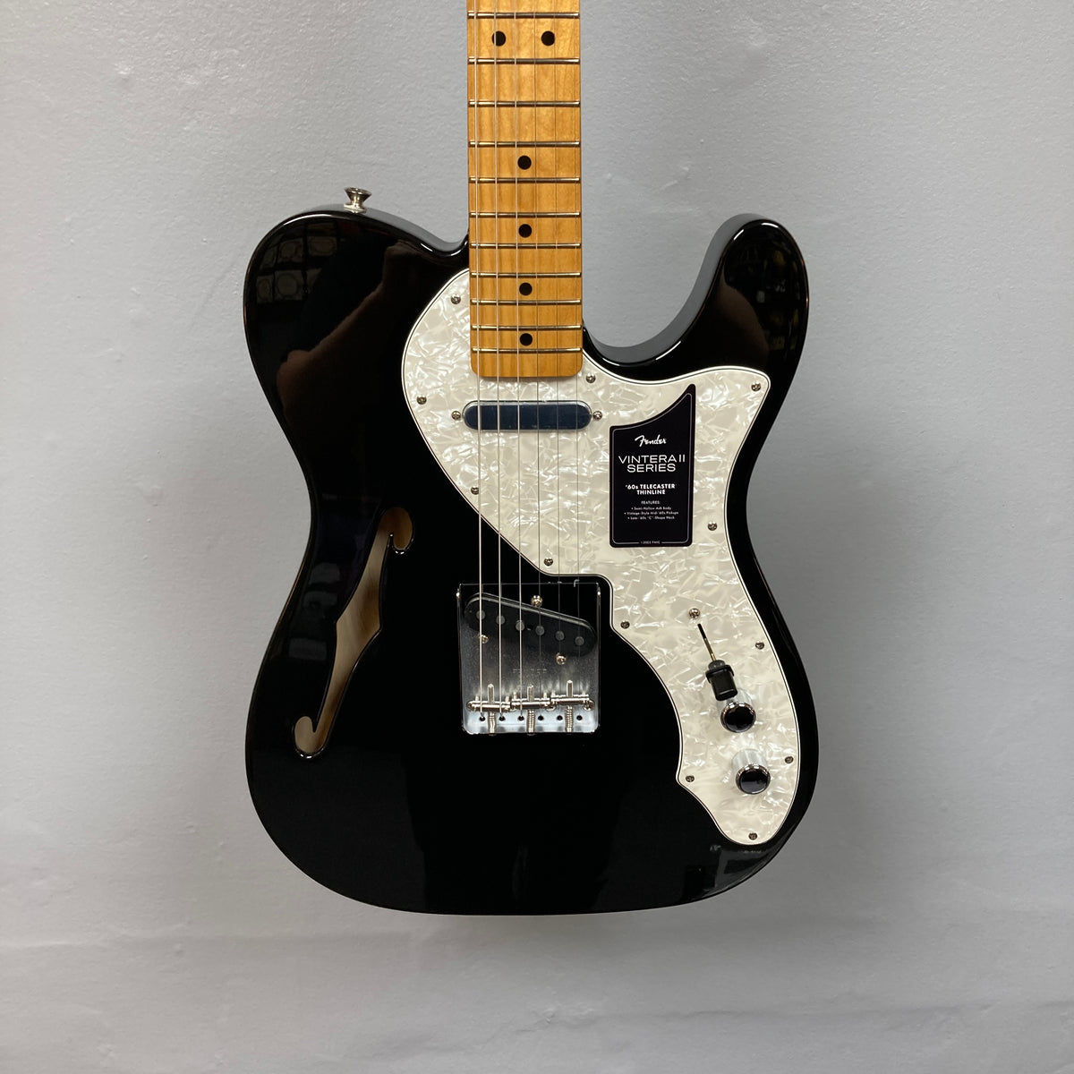 A used Fender Vintera II ’60s Telecaster Thinline in black, featuring a semi-hollow ash body, white pearloid pickguard, maple neck, three control knobs, and vintage-style Tele pickups from Fender, is hanging on a light gray wall.