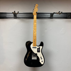 A used Fender Vintera II ’60s Telecaster Thinline electric guitar in black, featuring a semi-hollow ash body, white pickguard, and maple neck, hangs on a wall-mounted rack against a plain white background, displaying its classic vintage-style Tele pickups and iconic Fender design.