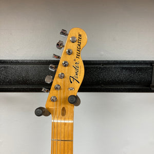 A Fender Vintera II ’60s Telecaster Thinline Black Used guitar headstock, complete with tuning pegs, the Fender logo, and a portion of the neck, is visible against a white wall and black horizontal rack. This model by Fender features vintage-style Tele pickups and a semi-hollow ash body for classic tone and resonance.