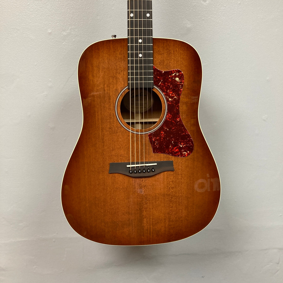 A Godin Century Maho Rustic Burst EQ SF solid wood guitar with a red tortoiseshell pickguard hangs on a plain light-colored wall.