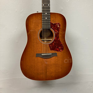 A Godin Century Maho Rustic Burst EQ SF solid wood guitar with a red tortoiseshell pickguard hangs on a plain light-colored wall.