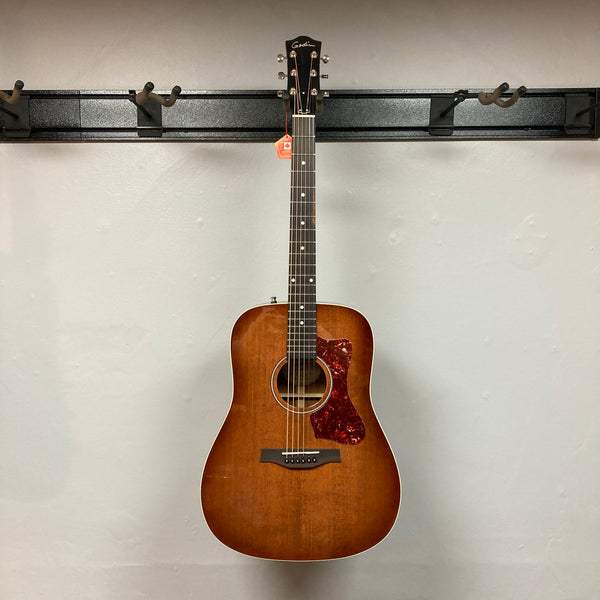 A Godin Century Maho Rustic Burst EQ SF solid wood guitar with a red tortoiseshell pickguard hangs on a plain light-colored wall.