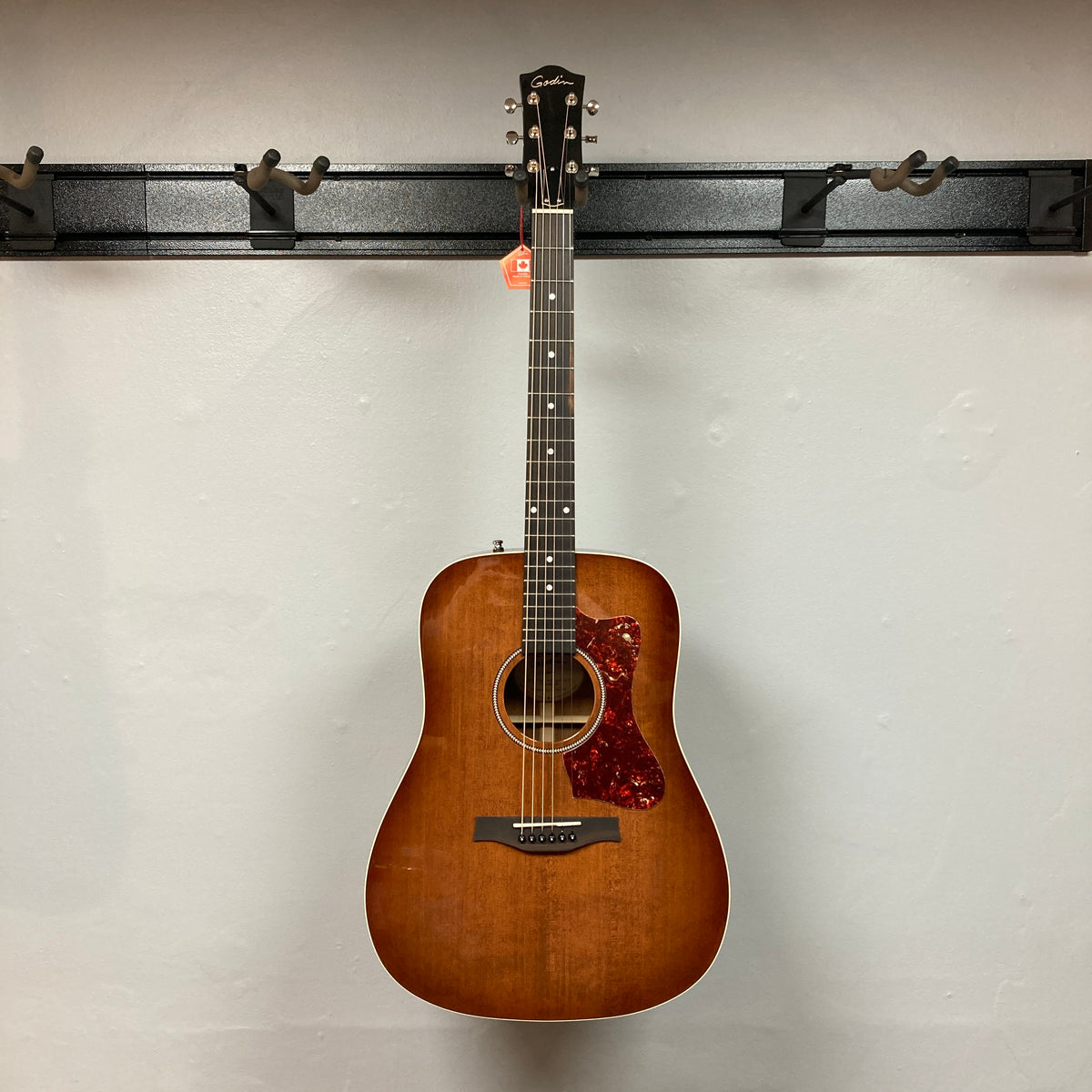 A Godin Century Maho Rustic Burst EQ SF by Godin, a dreadnought acoustic-electric guitar with a brown sunburst finish, hangs on a wall-mounted rack. The guitar features six strings, a pickguard, and is crafted from solid wood—blending classic aesthetics with modern electronics for versatile performance.
