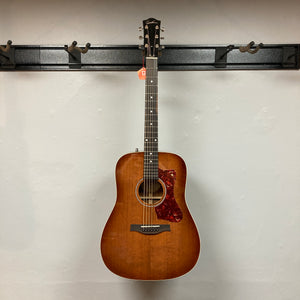 A Godin Century Maho Rustic Burst EQ SF by Godin, a dreadnought acoustic-electric guitar with a brown sunburst finish, hangs on a wall-mounted rack. The guitar features six strings, a pickguard, and is crafted from solid wood—blending classic aesthetics with modern electronics for versatile performance.
