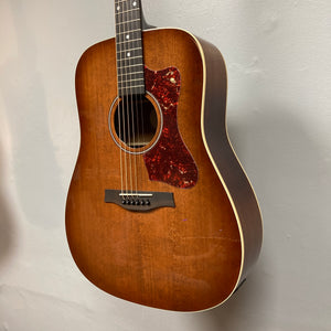 A Godin Century Maho Rustic Burst EQ SF acoustic guitar with a tortoiseshell-patterned pickguard is hanging on a light gray wall.
