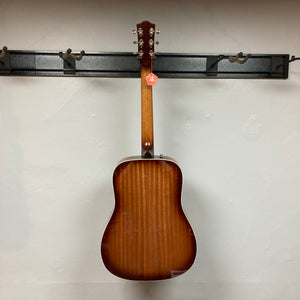 A Godin Century Maho Rustic Burst EQ SF acoustic guitar in brown, seen from the back, hangs on a black wall-mounted rack against a plain white wall.