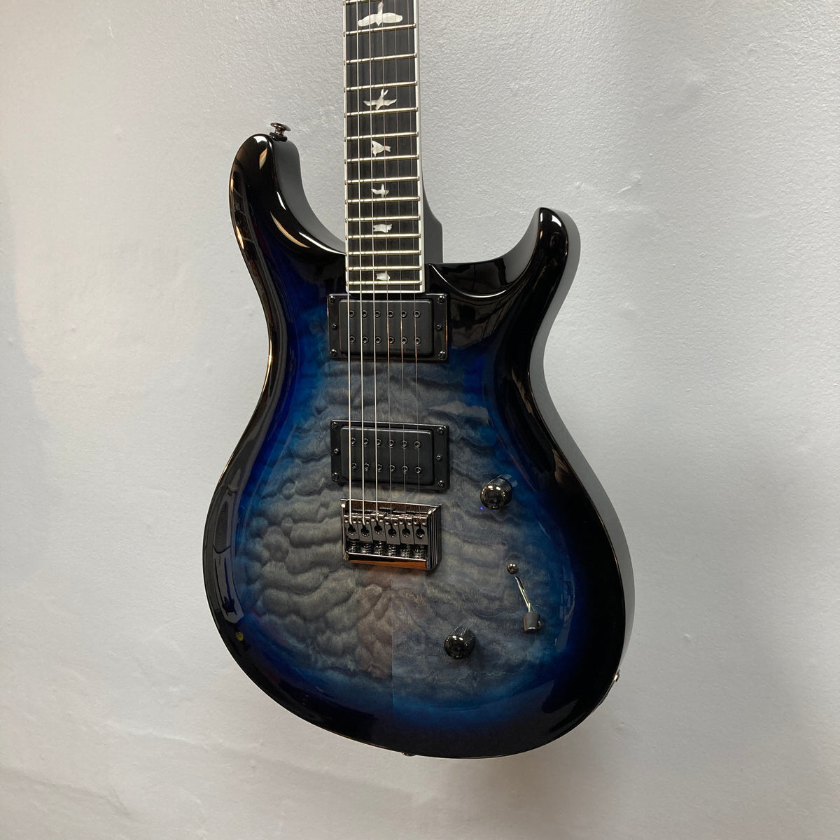 PRS SE Mark Holcomb - Holcomb Blue Burst w/Gigbag Used Consign by PRS, featuring quilted maple top, Seymour Duncan humbuckers, three knobs, and bird inlays, displayed against a white wall.